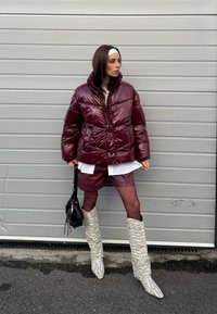 Femme portant une doudoune brillante bordeaux, un short assorti, des collants noirs à pois, des bottes blanches en tricot, un bandeau blanc, et tenant un sac à main noir.