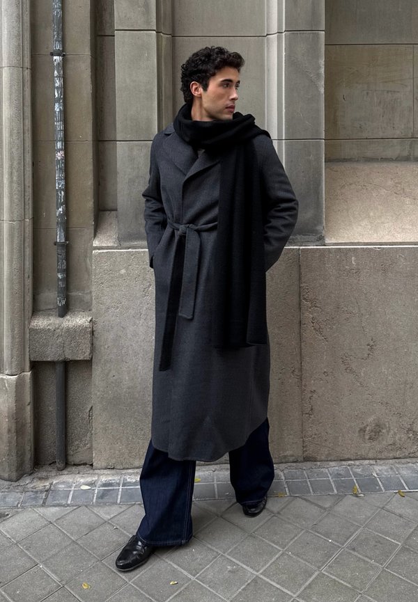 Homme debout contre un mur de pierre, portant un long manteau sombre, une écharpe noire, un pantalon large sombre et des chaussures noires, regardant vers sa droite.