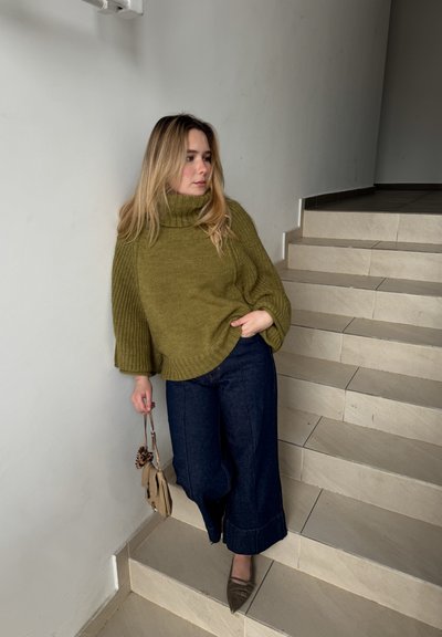 Suéter de gran tamaño en verde oliva con cuello de tortuga y tejido texturizado, combinado con pantalones de mezclilla anchos de color oscuro. Un bolso beige y zapatos topo completan el look.