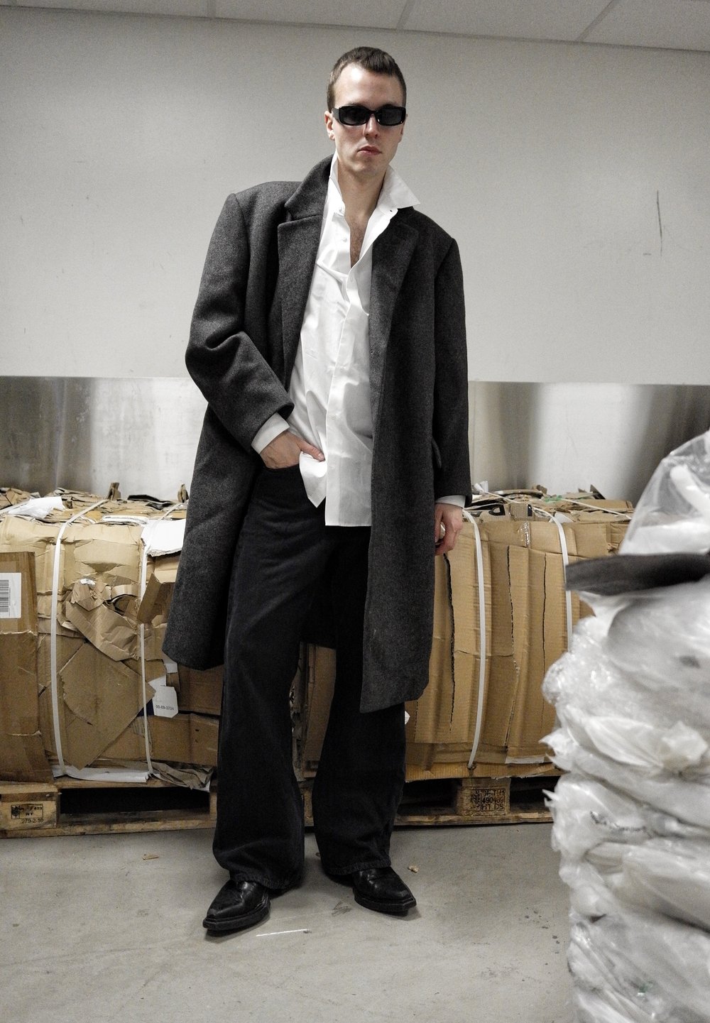 Homme portant un manteau noir surdimensionné, une chemise blanche, un pantalon large noir et des lunettes de soleil se tient dans une zone de stockage industrielle avec des balles de cartons derrière.
