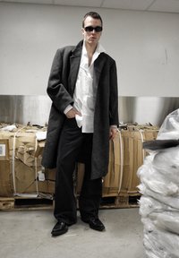 Homme portant un manteau noir surdimensionné, une chemise blanche, un pantalon large noir et des lunettes de soleil se tient dans une zone de stockage industrielle avec des balles de cartons derrière.
