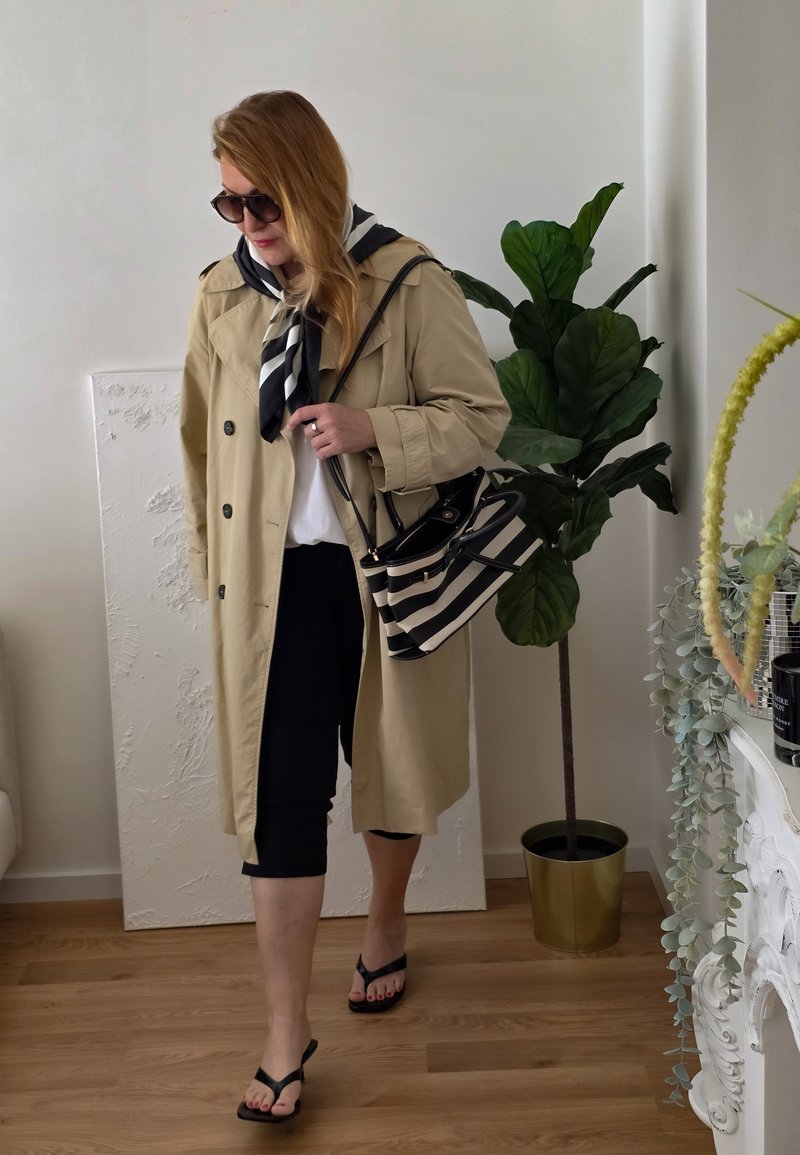 Kvinne iført beige trenchcoat, svarte capribukser, flip-flops, solbriller, bærer stripet håndveske og svart-hvitt skjerf innendørs nær en plante.