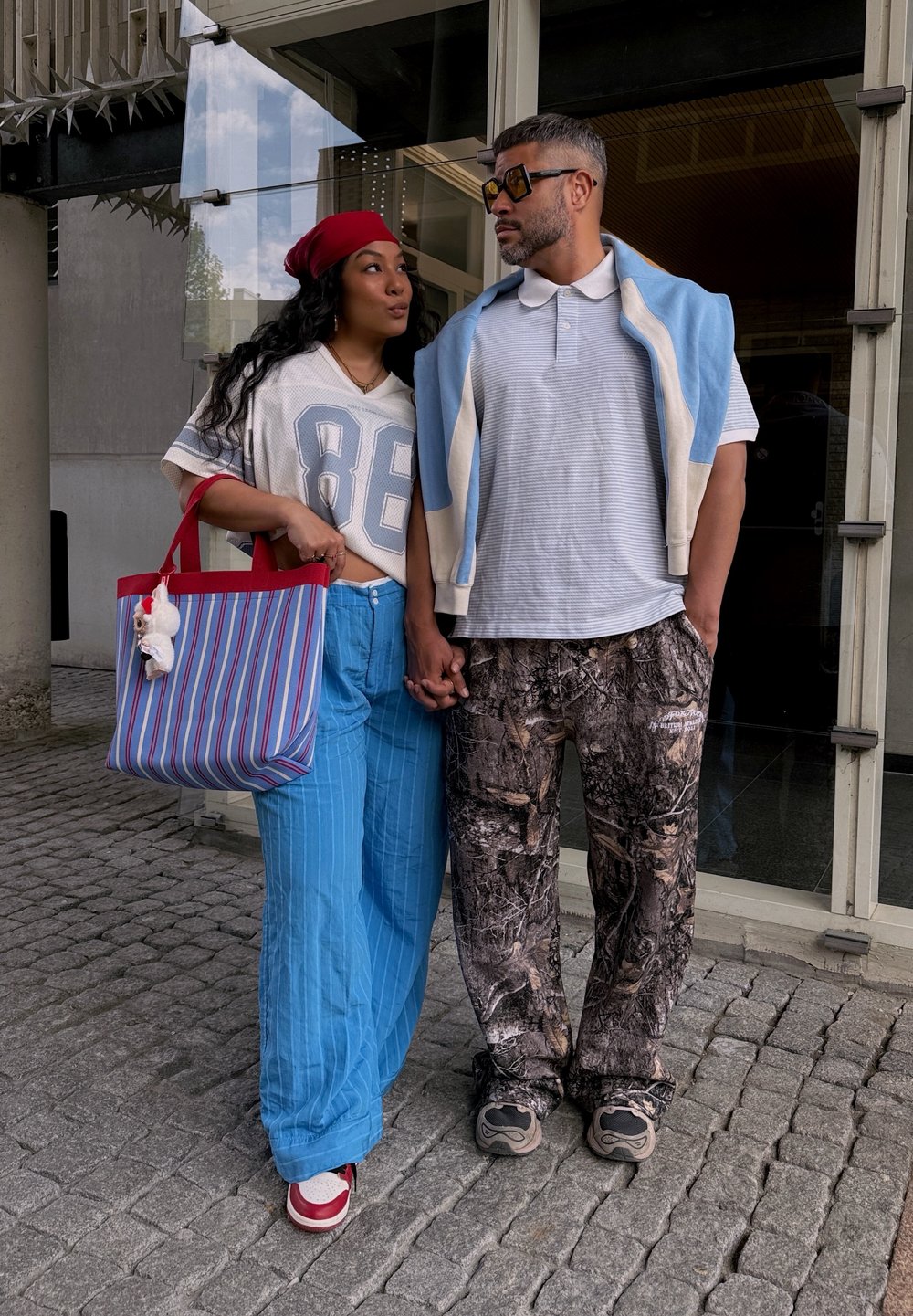 Δύο άτομα στέκονται δίπλα-δίπλα. Ο ένας φορά μια λευκή ριγέ μπλούζα, μπλε πουλονγκ και παραλλαγής παντελόνι. Ο άλλος φορά μια διαφανή μπλούζα και μπλε ριγέ παντελόνι. Μια ριγέ τσάντα ώμου κρέμεται από τον έναν ώμο, με ένα πλαστικό γούρι.