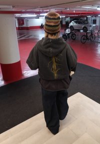 Felpa marrone con cappuccio e motivi gialli, pantaloni scuri e un cappello lavorato a maglia con strisce multicolori. Ambientazione in un parcheggio rosso e grigio.