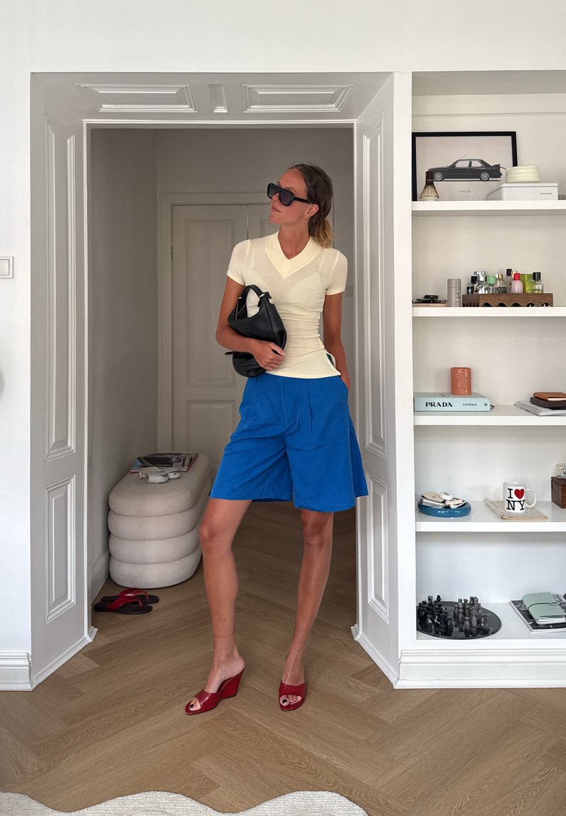 Une femme se tient dans une embrasure de porte, vêtue d’un haut blanc à manches courtes, d’un short bleu, de chaussures compensées rouges, de lunettes de soleil noires et tenant un sac à main noir.