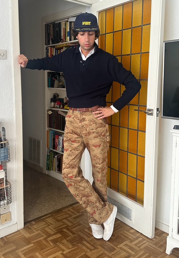 Hombre con un suéter negro, pantalones beige estampados, zapatillas blancas y un sombrero azul marino, de pie con una mano en la cadera y la otra en el marco de la puerta.