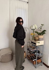 Femme portant des lunettes de soleil et une veste noire se tient près d'une étagère avec des plantes, des magazines, une lampe et un baby-foot de table à l'intérieur.