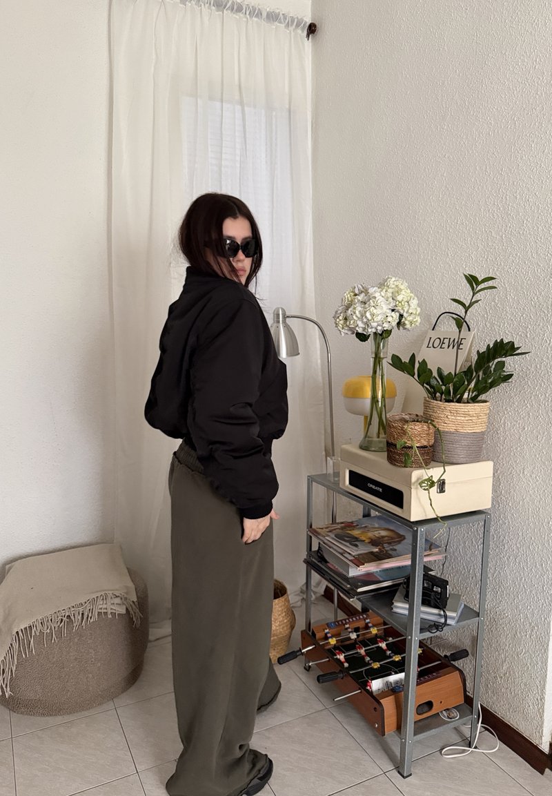 Woman wearing sunglasses and black jacket stands near shelf with plants, magazines, a lamp, and a tabletop foosball game indoors.