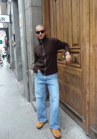 Suéter marrón de canalé con cremallera, jeans anchos de color azul claro, zapatos marrones sin cordones. De pie junto a una puerta de madera con herrajes de bronce redondeados.