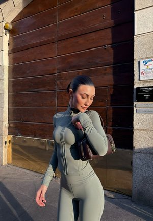 Mujer con ropa deportiva ajustada gris posa al aire libre bajo la luz del sol, sosteniendo un pequeño bolso marrón sobre el hombro frente a una puerta de madera.