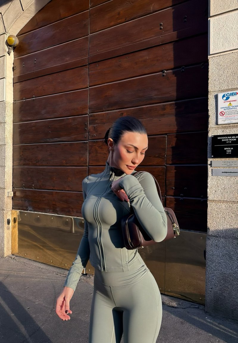 Femme en tenue de sport gris ajustée, debout à l'extérieur sous le soleil, tenant un petit sac à main marron posé sur son épaule devant une porte en bois.