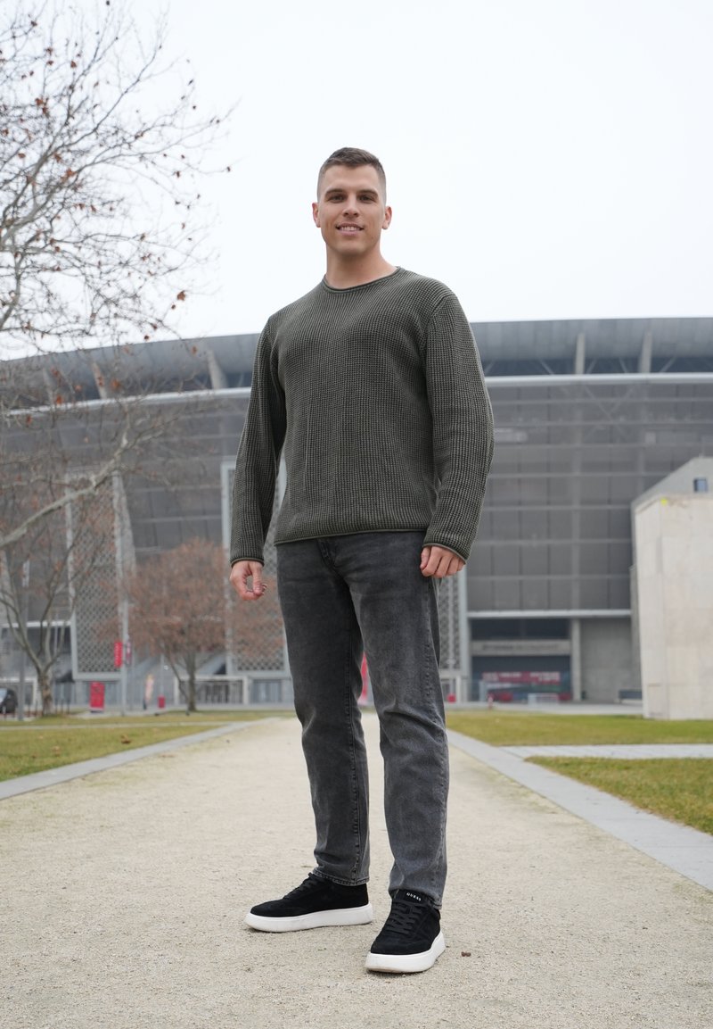 Uomo che indossa un maglione verde scuro a trama, jeans grigi e sneakers nere con suola bianca, in piedi su un sentiero con uno stadio sullo sfondo.