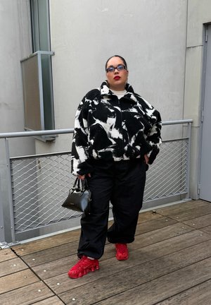 Persona con chaqueta de estampado blanco y negro, pantalones negros, zapatillas rojas y gafas de sol, sosteniendo un pequeño bolso negro en una plataforma de madera.