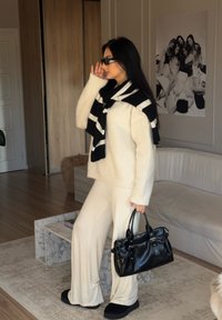 Cream textured jumper, black and white striped scarf, cream wide-leg trousers, black handbag, and black slippers in an indoor setting.
