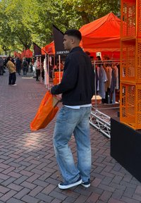 En person holder en orange tote bag, iklædt en sort jakke og lyseblå jeans, ved et marked med orange markiser og tøjstativer.