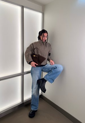 Mujer con chaqueta de cuadros y vaqueros azules levanta una pierna, sosteniendo un bolso triangular marrón, de pie frente a una pared blanca con paneles.