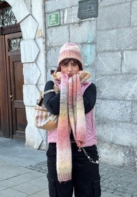 Bonnet et écharpe rayés en rose et beige tricotés, gilet rose porté sur un top noir, pantalon cargo noir, et sac à main rayé avec un accent chaîne.