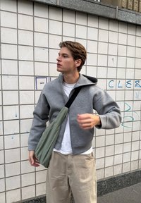 Grau zip-up Hoodie mit großer Kapuze, über einem weißen T-Shirt getragen, kombiniert mit beigen, lässig sitzenden Hosen und einer grünen Umhängetasche.