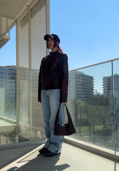 Chaqueta de cuero marrón, jeans de color azul claro y zapatos negros. Lleva un bolso de mano morado oscuro. De pie contra un cielo brillante y despejado.