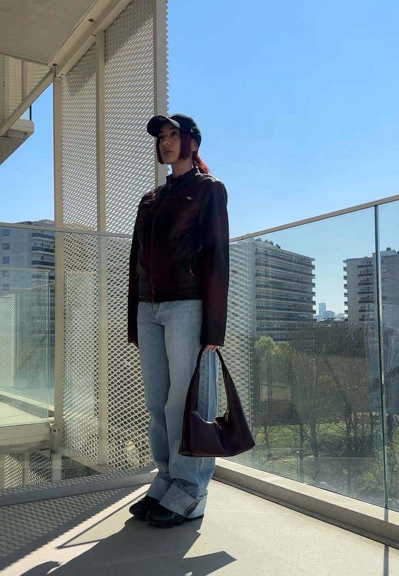 Jaqueta de couro castanha, jeans azul claro e sapatos pretos. A segurar uma bolsa roxa escura. Em pé contra um céu brilhante e limpo.