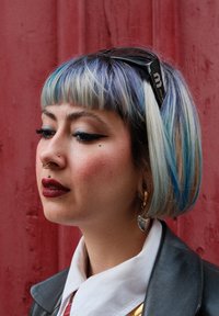 Coupe bob courte avec des couleurs bleu et blond dégradées. Présente un eye-liner audacieux, un piercing au septum, des boucles d'oreilles en or et une veste en cuir.