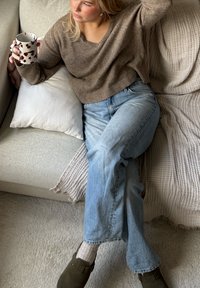 Brązowy sweter dzianinowy, jasnoniebieskie dżinsy proste, szare skarpetki i ciemne klapki. Trzyma białą filiżankę w burgundowe plamki, siedząc na sofie.