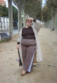 Trench coat beige sopra un top marrone, pantaloni sportivi marroni con strisce bianche e scarpe con tacco marroni. Porta una borsa scura e si trova su un sentiero.