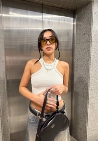 Balta raukšlėta halterio tipo palaidinė, džinsinė sijonas, juoda rankinė ir geltonai dažyti stačiakampiai akiniai. Sidabro papuošalai ir tekstūruota akmeninė fono.