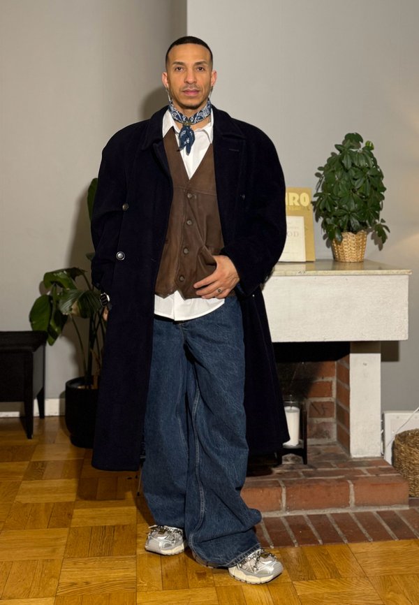Homme portant un pardessus sombre, un gilet marron, une chemise blanche, un jean bleu, des baskets et un foulard bandana bleu, debout à l'intérieur près d'une cheminée.