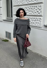 Kvinna med lockigt svart hår, klädd i en grå avskuren tröja, svart satinkjol, grå sneakers och bär en vinröd handväska, går utomhus.