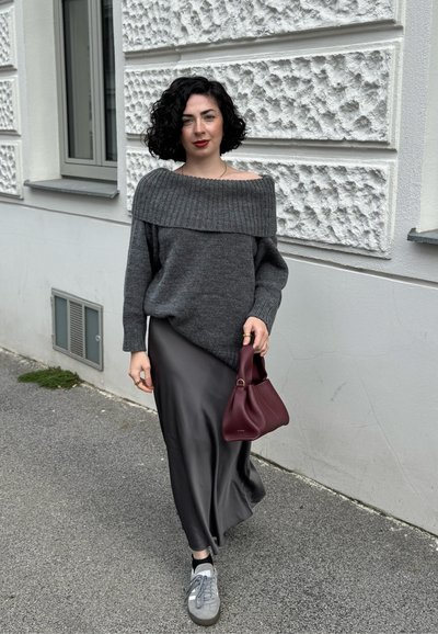 Mujer con cabello negro rizado que lleva un suéter gris off-shoulder, falda de satén negra, zapatillas grises y sosteniendo un bolso burdeos caminando al aire libre.