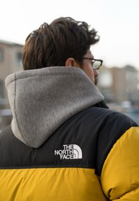 Doudoune à design bicolore ; noir en haut, jaune vif en dessous, et capuche grise. Logo brodé sur l'épaule.