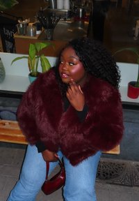 Γυναίκα με σγουρά μαλλιά που φοράει μπορντό γούνινο παλτό και μπλε τζιν κάθεται σε ένα παγκάκι, κρατώντας μια τσάντα σε ταιριαστή μπορντό απόχρωση και κοιτάζει προς τα πλάγια.