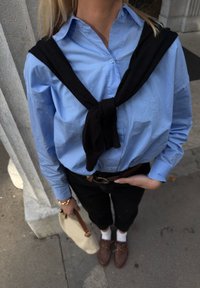 Persona vestita con una camicia azzurro chiaro con un maglione nero annodato sulle spalle, pantaloni neri, scarpe marroni, calzini bianchi e che tiene una borsa beige.