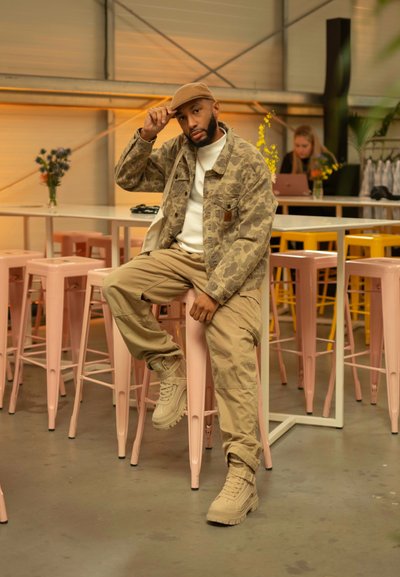 Chaqueta con patrón de camuflaje, sudadera blanca, pantalones caqui y botas de color beige claro. Sentado en un taburete de metal rosa en un entorno brillante.