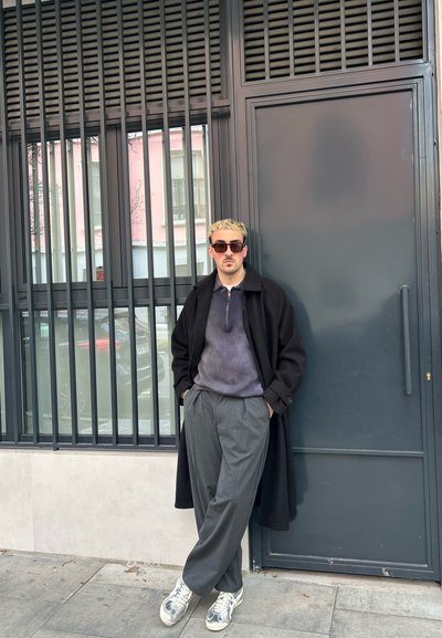 Abrigo negro, suéter gris con cremallera, pantalones grises con pliegues y zapatillas plateadas reflectantes. De pie contra una pared gris con barras verticales.