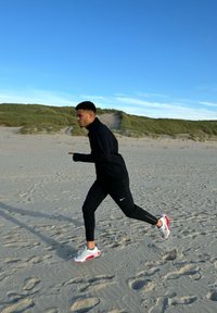 Must musternäidud ja püksid, valged ja punased jooksujalanõud, joostes liivarannal roheliste düünide ja sinise taeva taustal.