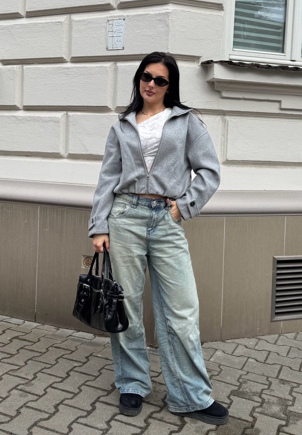 Γυναίκα που φοράει γκρι ζακέτα με φερμουάρ, λευκό τοπ, τζιν παντελόνι με φαρδιά πόδια, μαύρα παπούτσια και γυαλιά ηλίου, κρατώντας μαύρη τσάντα στην πεζοδρόμιο της πόλης.