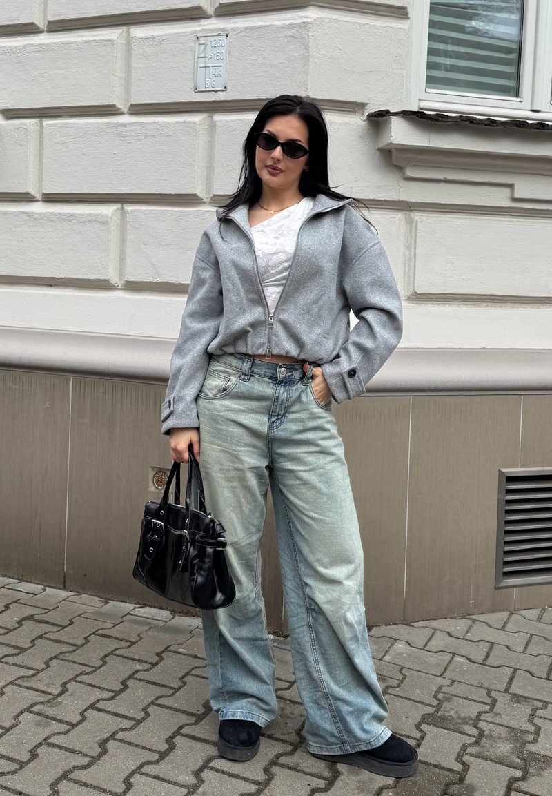 Femme portant une veste grise zippée, un haut blanc, un jean à jambes larges, des chaussures noires et des lunettes de soleil, tenant un sac à main noir sur un trottoir en ville.
