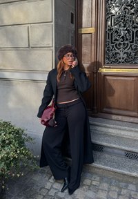 Svart lång kappa, brun skjorta och byxor, kombinerat med en vinröd handväska och en brun pälsmössa. Står vid en texturerad stenvägg och en dörr.