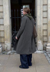 Pikk, hall ruuduline mantel rohelise kapuutsiga kihiga all, siniste teksade ja mustade kingadega. Seisab kivimüüri vastu.