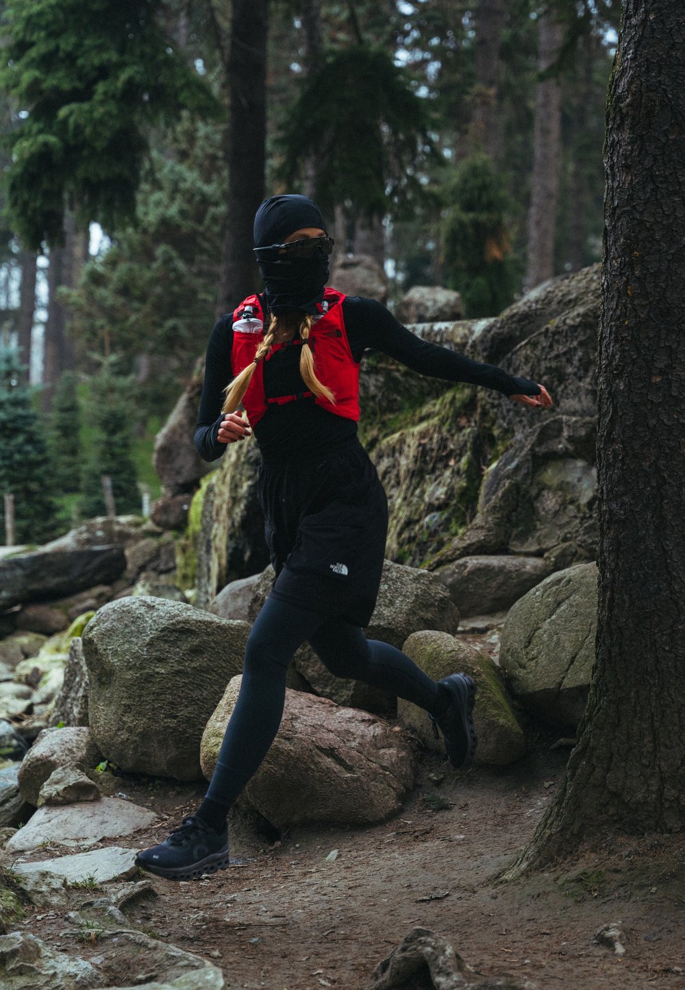 Gilet de course rouge porté sur un haut à manches longues noir, short noir et leggings. Scène en plein air avec un terrain rocheux et des arbres en arrière-plan.