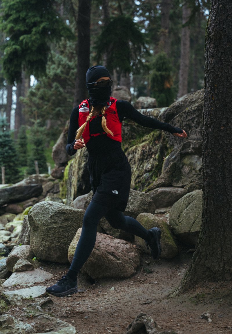 Gilet de course rouge porté sur un haut à manches longues noir, short noir et leggings. Scène en plein air avec un terrain rocheux et des arbres en arrière-plan.