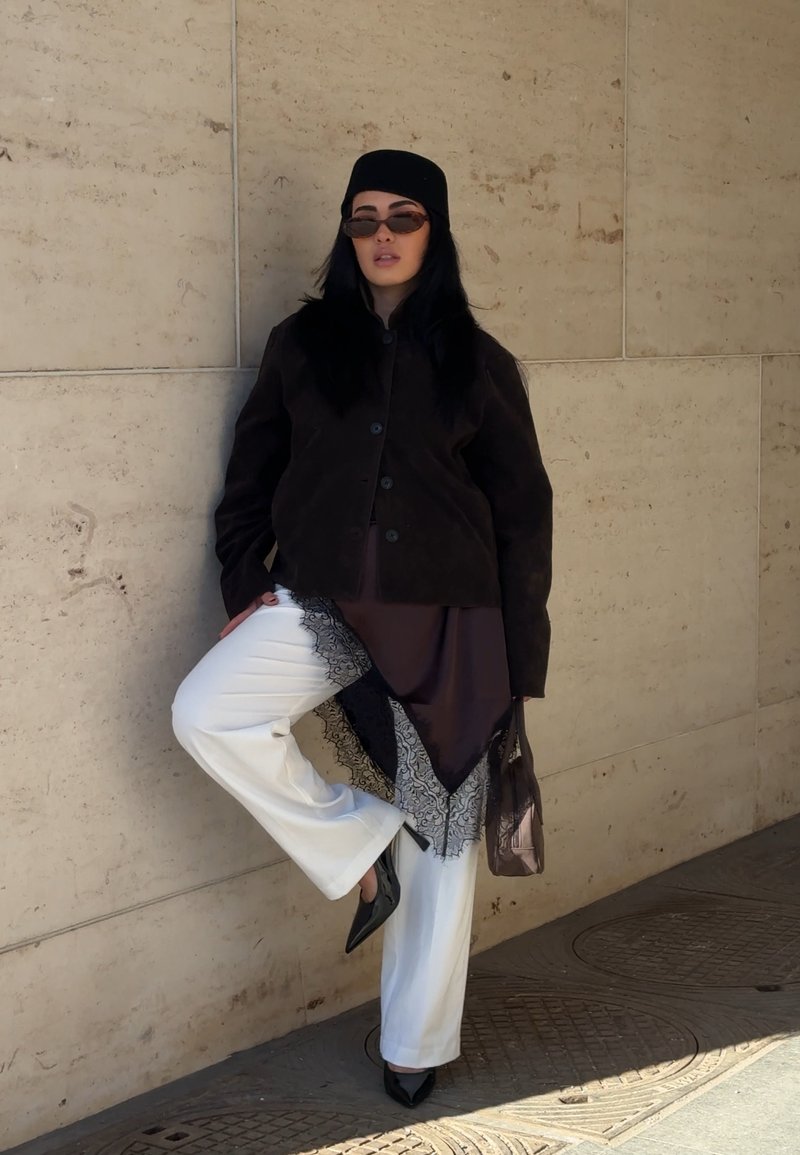 Woman wearing black jacket, hat, sunglasses, white pants, and black heels poses against beige stone wall with one leg raised.