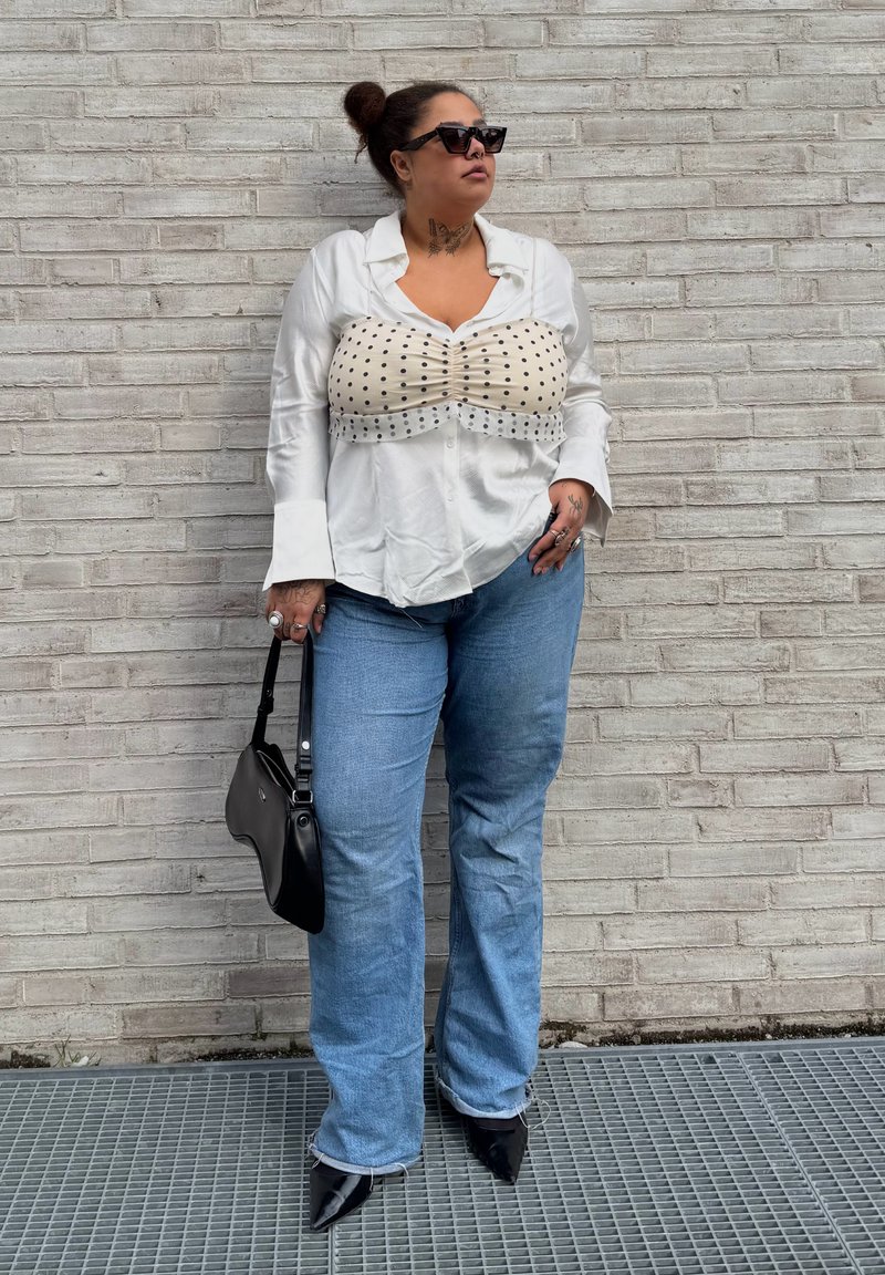 Žena s okuliarmi na slnko a vo vlasoch vrkoč, oblečená v bielej košeli s vrchným dielom s bodkami, modré džínsy, čierne špicaté topánky, drží čiernu kabelku pri tehlovej stene.
