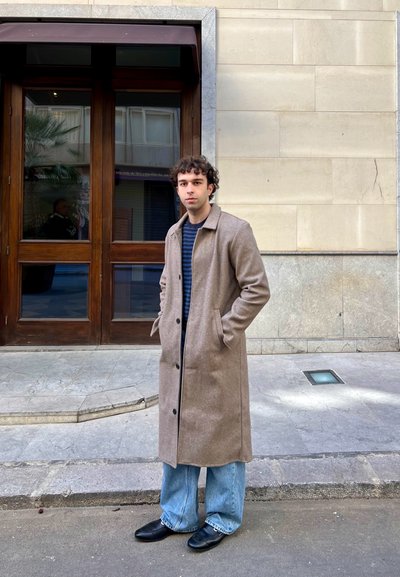 Abrigo largo de mezcla de lana marrón, camisa de rayas azul marino, jeans de pierna ancha azul claro y zapatos negros, posando al aire libre cerca de puertas de madera y pavimento de piedra.