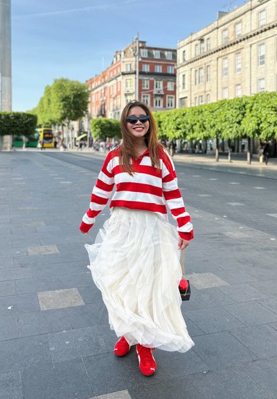 Top de manga larga a rayas rojas y blancas, falda blanca fluida con volantes y zapatillas rojas. Accesorio: pequeña bolsa negra cruzada. Entorno urbano.