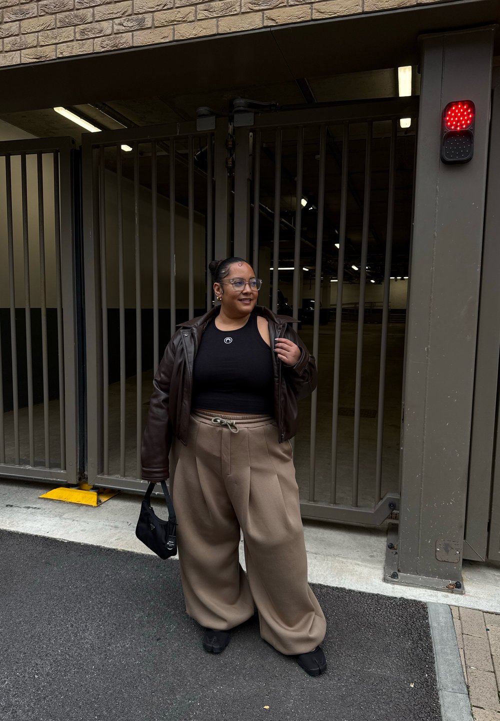 Veste en cuir marron sur un débardeur noir court, pantalon large beige, sac noir, debout à une entrée avec portail dans un environnement de garage.