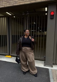 Veste en cuir marron sur un débardeur noir court, pantalon large beige, sac noir, debout à une entrée avec portail dans un environnement de garage.