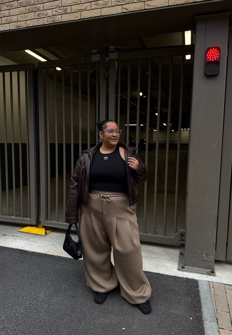 Brown leather jacket over a black cropped tank, beige wide-leg pants, black bag, standing at a gated entrance in a garage setting.